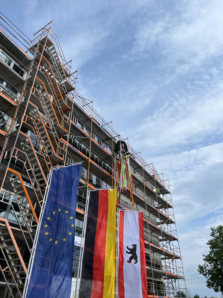Aufnahme des Schulgebäudes von unten. Man sieht den blauen Himmel. Am Baugerüst hängt oben der Richtkranz, darunter die Flaggen von Europa, Deutschland und Berlin.