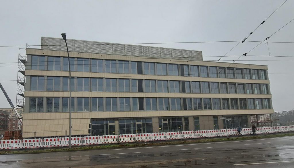 Das Schulgebäude wurde von der gegenüberliegenden Straße fotografiert. Die Außenfassade ist nun zu weiten Teilen fertiggestellt. Das Gebäude ist von außen beige, aber besteht vor allem zu großen Teilen aus Fensterfassade.